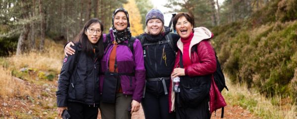 A group of four women ramblers stop on their walk and smile to the camera, with their arms around eath other. They have been walking together through a woodland path on a cold brisk day